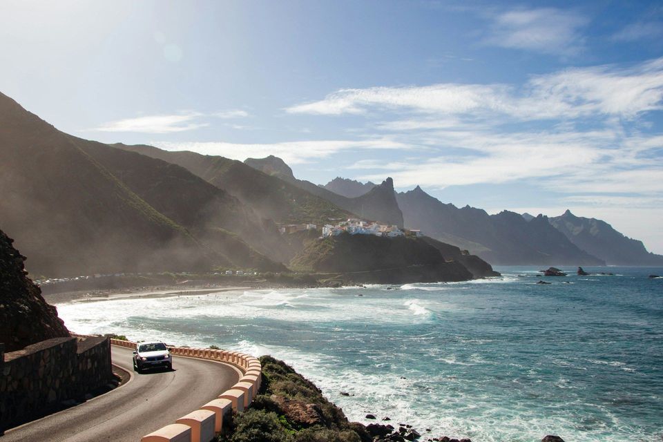 Un'auto percorre una strada tortuosa lungo una costa frastagliata, con montagne lontane e un piccolo villaggio sotto un cielo soleggiato.