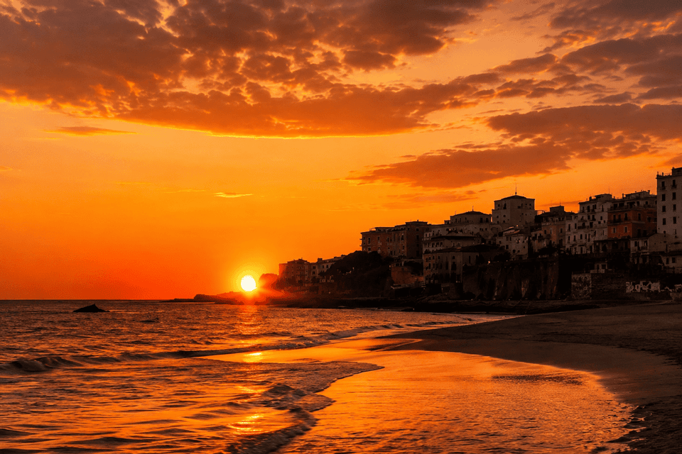 Un tramonto arancione si riflette sul mare e su una spiaggia di sabbia bagnata, con una città costiera stagliata su una scogliera sullo sfondo.