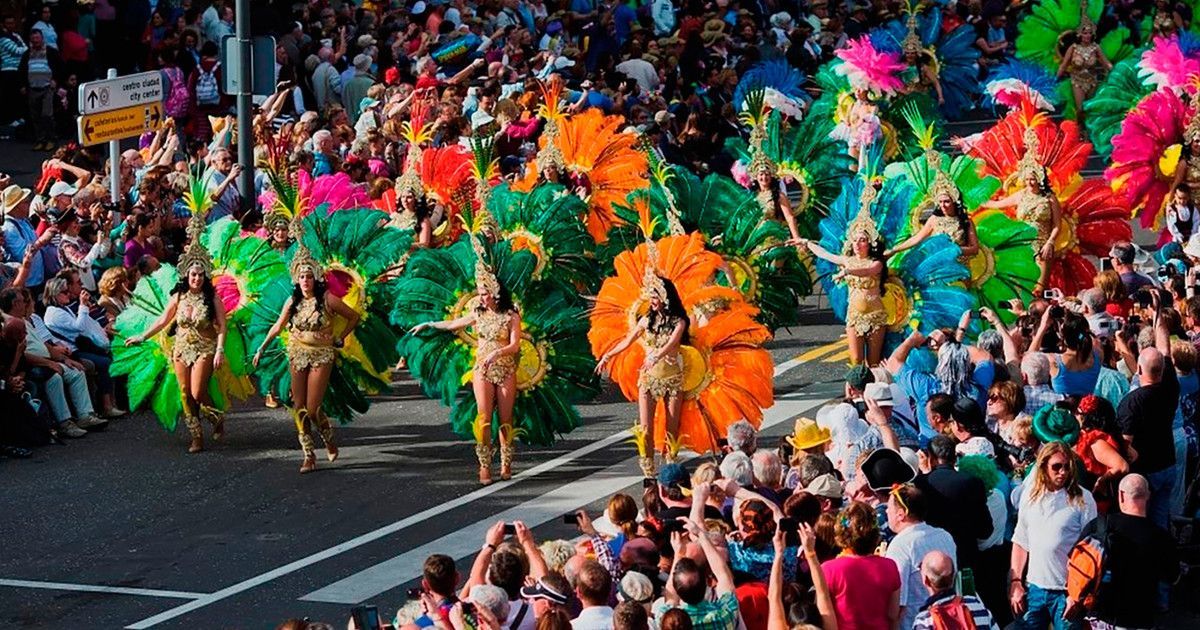 Danzatori in costumi colorati e piumati sfilano per una strada, osservati da una grande folla di spettatori.