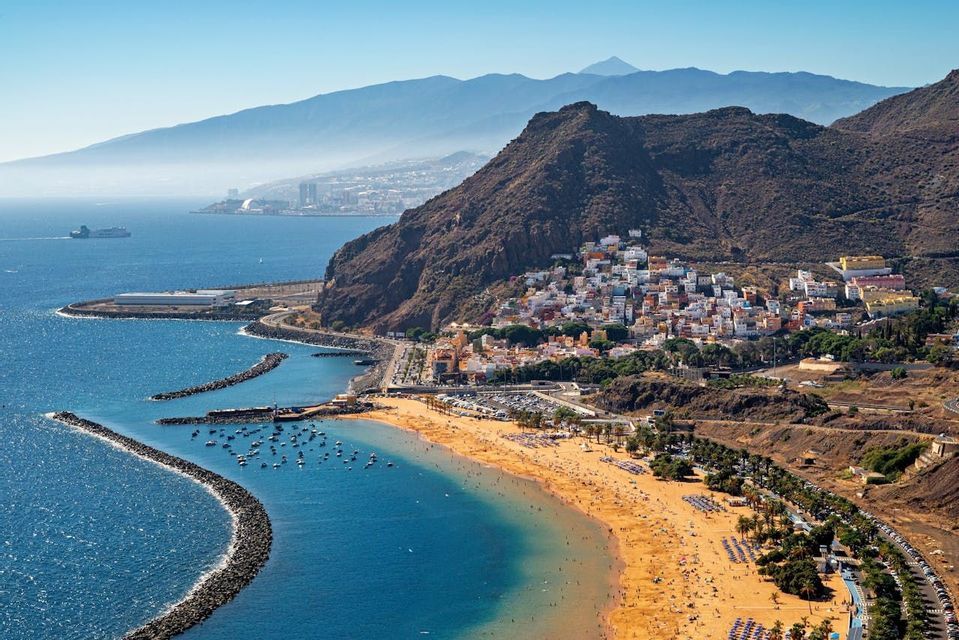 Una veduta aerea di una spiaggia affollata di sabbia dorata accanto a una località costiera, con montagne sullo sfondo sotto un cielo sereno.
