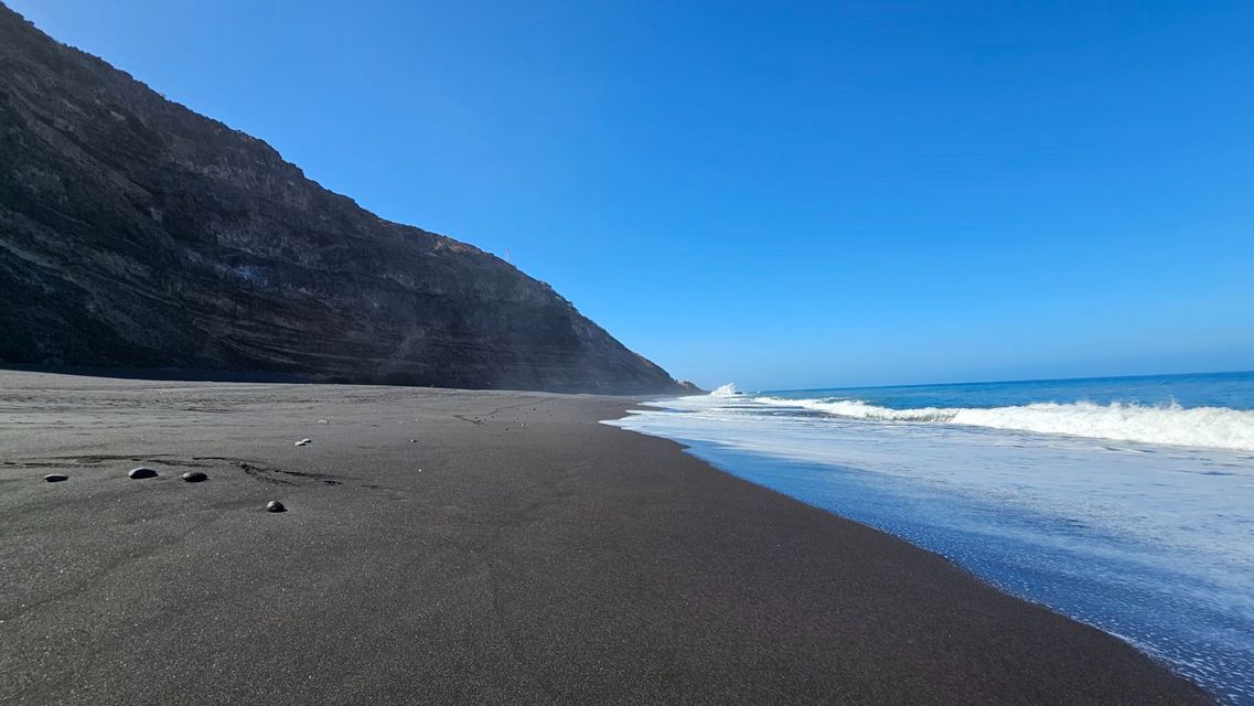 Weiße Wellen spülen an einen schwarzen Sandstrand neben einer steilen, felsigen Klippe unter klarem blauem Himmel.