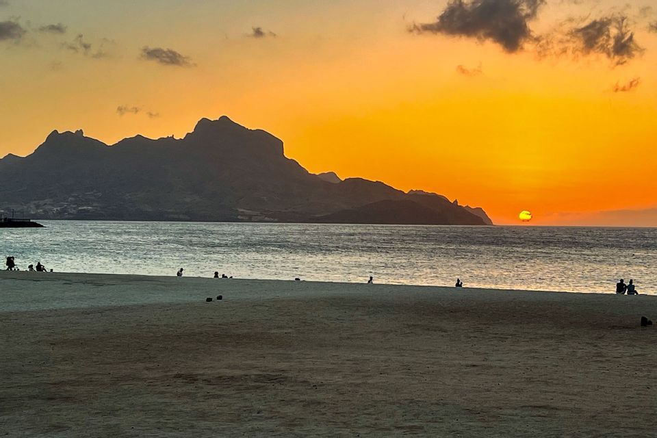 Menschen sitzen an einem Sandstrand und beobachten den Sonnenuntergang über dem Meer hinter einer schattenhaften Bergkette unter einem orangefarbenen Himmel.