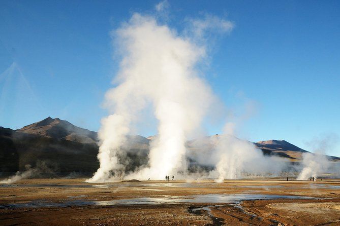 A WeRoad group trip observes large geysers erupting steam in a vast, arid valley with mountains in the background.