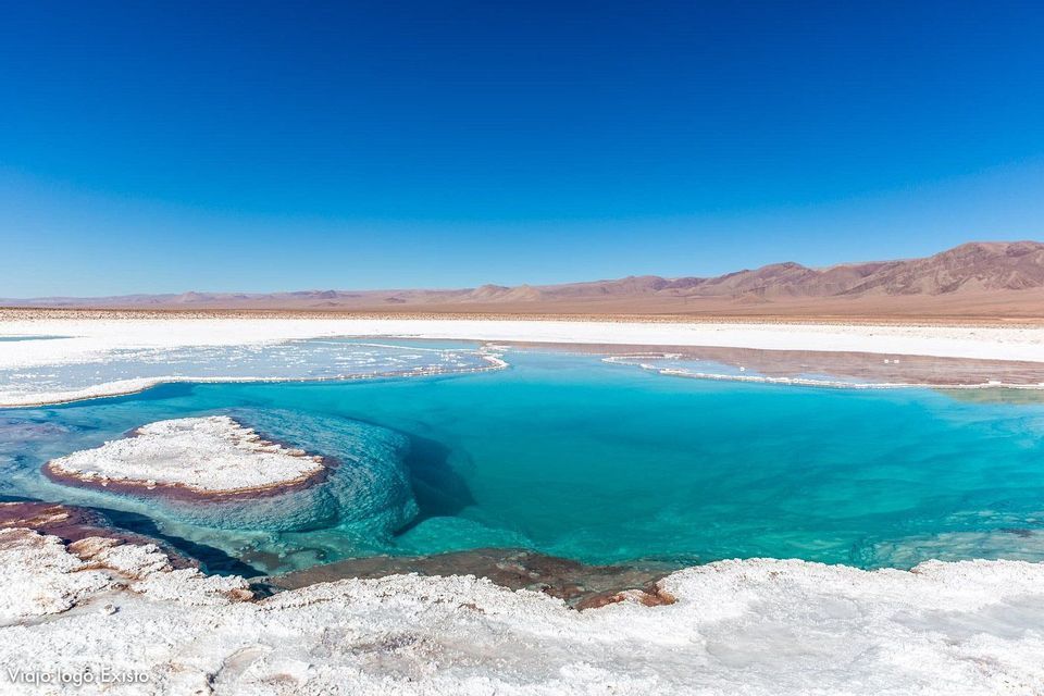 A pool of clear turquoise water surrounded by white salt flats, with distant mountains under a blue sky.