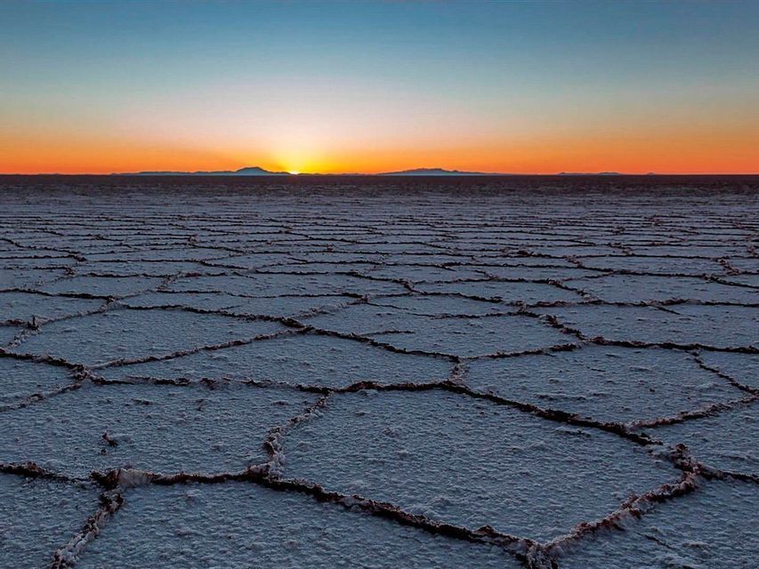 The sun sets on the horizon over a vast salt flat with a cracked, geometric pattern on the ground.