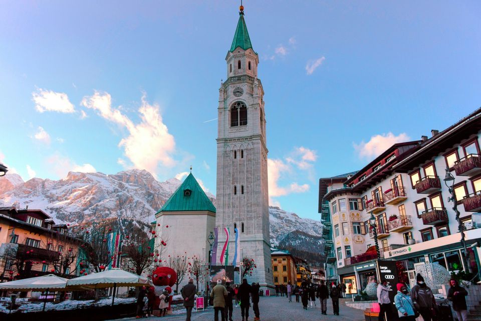 Un alto campanile in pietra con una guglia verde si staglia su una piazza animata da persone, con lo sfondo di montagne innevate.