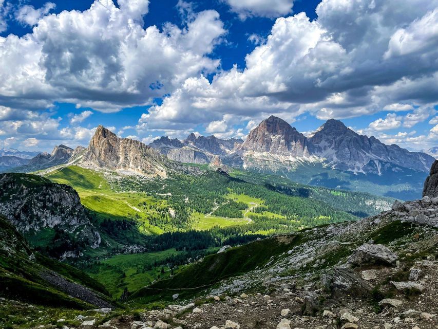 Una vista panoramica di una catena montuosa rocciosa con valli verdi e foreste sotto un cielo blu con nuvole bianche e gonfie.