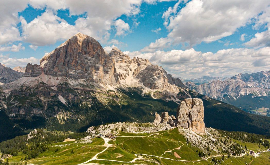 Una vista panoramica di una catena montuosa rocciosa, con cime prominenti, colline verdi con sentieri tortuosi e un cielo parzialmente nuvoloso.