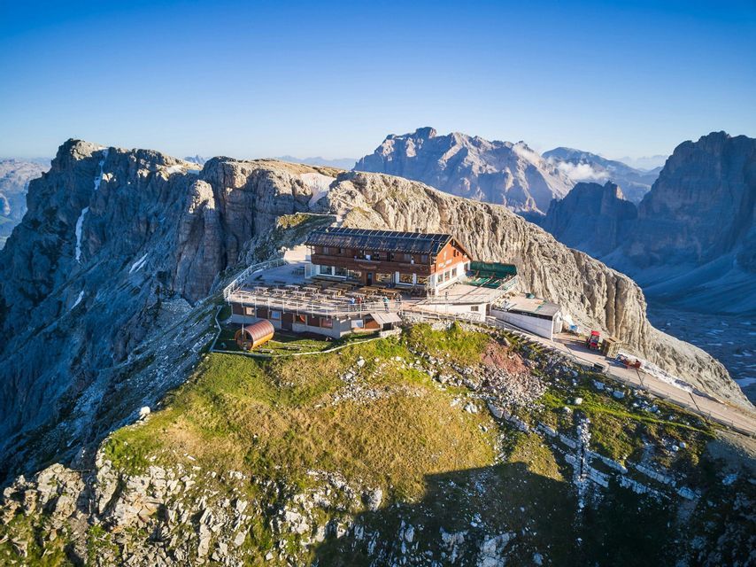Un rifugio di montagna con un'ampia terrazza si trova su una cima erbosa, circondato da vette rocciose sotto un cielo azzurro e limpido.