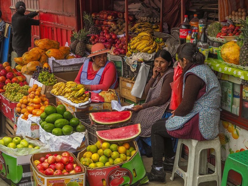 Three women vendors sit and talk at a colorful outdoor market stall filled with a variety of fresh fruits.
