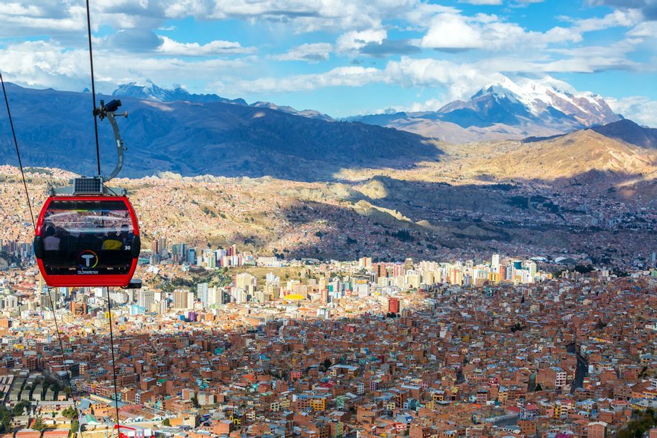 Eine rote Seilbahn gleitet über eine weitläufige Stadt in einem Tal, mit großen schneebedeckten Bergen im Hintergrund.