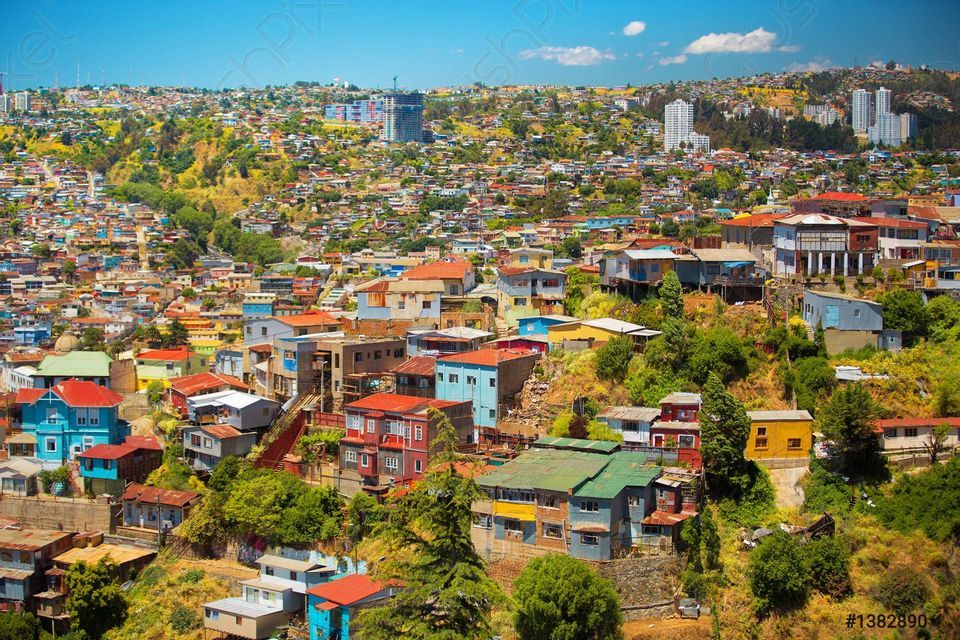 A dense cityscape of colorful houses and buildings covering a sprawling green hillside under a clear blue sky.