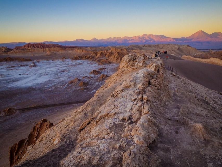 A WeRoad group trip stands on a rocky ridge overlooking a vast desert valley with distant mountains at sunset.