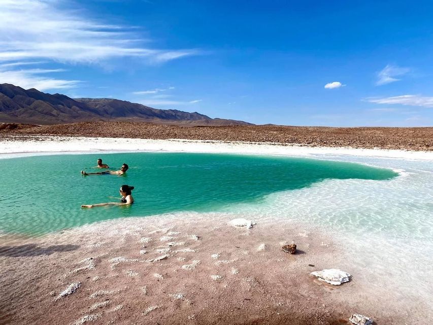 A WeRoad group trip of three people floating in a turquoise salt lake, surrounded by a desert landscape and mountains.