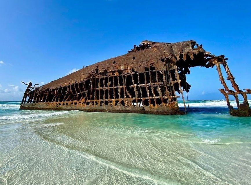 Un gran naufragio oxidado descansa en una playa de arena, parcialmente sumergido en aguas cristalinas de color turquesa bajo un cielo azul.