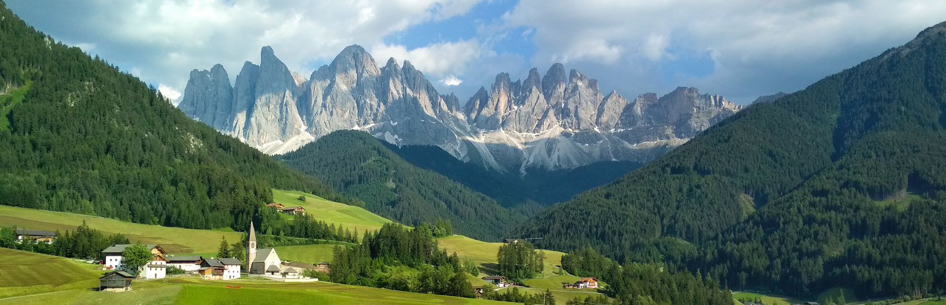 Un pequeño pueblo con una iglesia se asienta en un valle verde al pie de colinas boscosas y una sierra montañosa escarpada y rocosa.