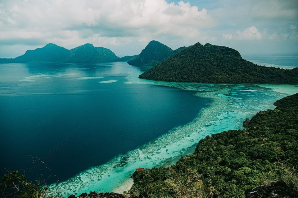 A high-angle view of lush, forested islands surrounded by turquoise and deep blue water, with a visible coral reef along the coastline.