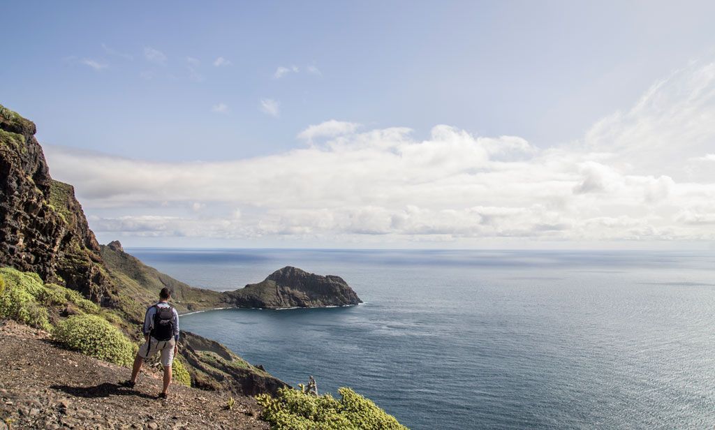 Un escursionista con uno zaino si trova su un sentiero costiero roccioso con vista sull'oceano e un promontorio distante sotto un cielo parzialmente nuvoloso.