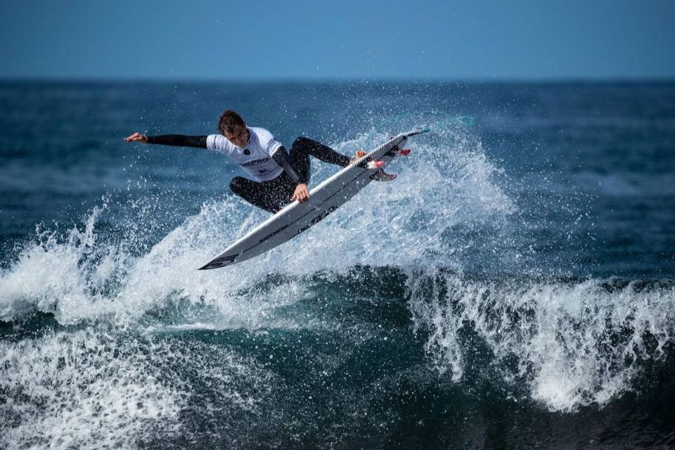 Un surfista con muta e maglia da competizione è in volo sulla sua tavola da surf, cavalcando la cresta di un'onda e creando spruzzi.