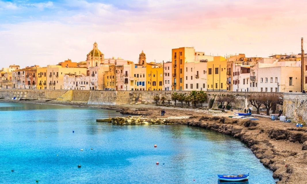 Edifici colorati di una città costiera si affacciano su una baia con acqua cristallina, con una piccola barca in primo piano durante l'alba.
