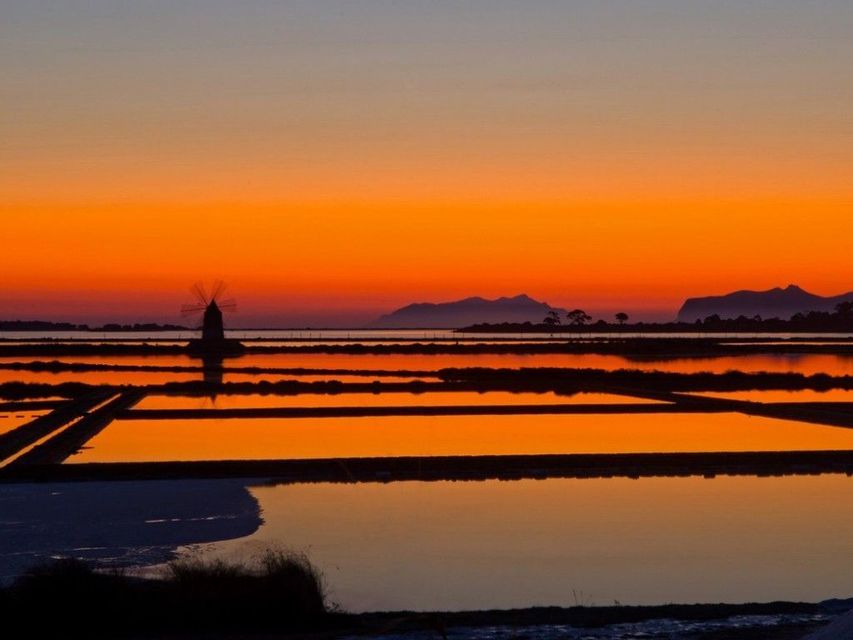 Una sagoma di mulino a vento si staglia sulle saline che riflettono un vibrante tramonto arancione, con le montagne in lontananza.