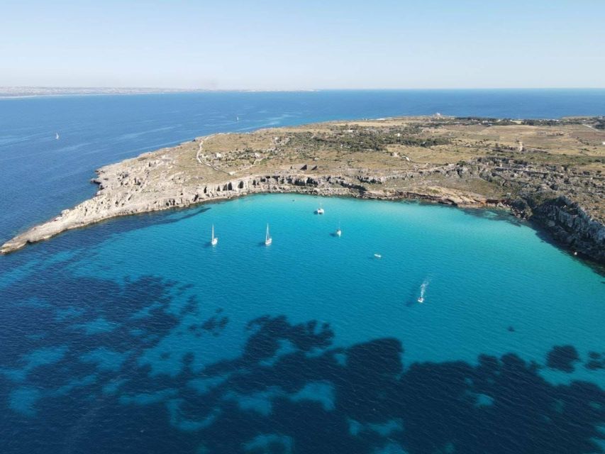 Una vista aerea di barche a vela in una baia turchese accanto a una penisola rocciosa sotto un cielo sereno.