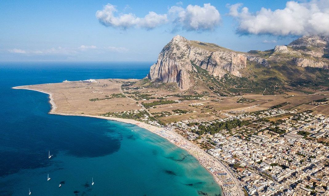Veduta aerea di una cittadina costiera e di una spiaggia affollata, lambite da acque turchesi, con una grande montagna rocciosa sullo sfondo.