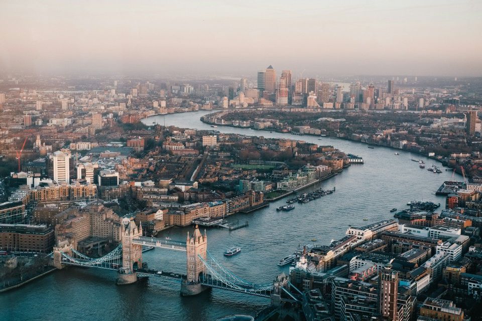 An aerial view of Tower Bridge and the River Thames winding through a dense cityscape towards a modern skyline under a hazy sky.
