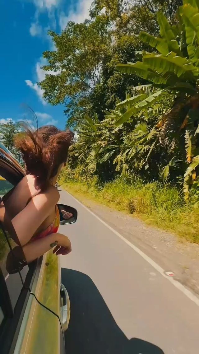 Una donna si sporge dal finestrino dell'auto con i capelli al vento, percorrendo una strada fiancheggiata da alberi verdi e lussureggianti.