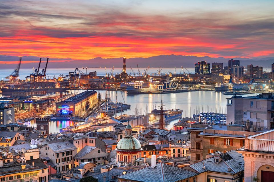 Una vista panoramica di una grande città portuale al tramonto, con edifici illuminati e barche nel porto sotto un cielo colorato.