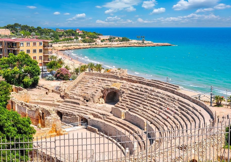 Un antico anfiteatro in pietra si affaccia su una spiaggia sabbiosa con acqua turchese e una città costiera sotto un cielo parzialmente nuvoloso.