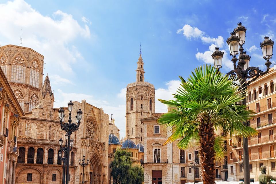 Una cattedrale di pietra con un alto campanile e cupole blu si erge in una piazza della città con una palma sotto un cielo azzurro.