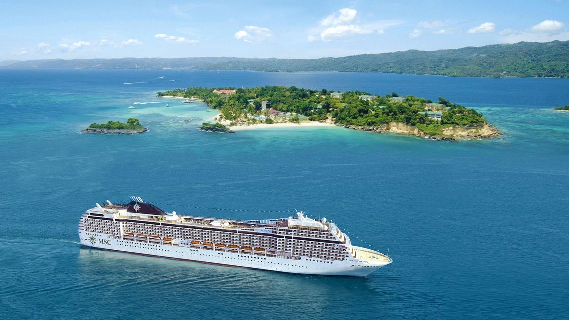 Una vista aerea di una grande nave da crociera bianca che naviga su un mare blu vicino a un'isola tropicale lussureggiante e verde.