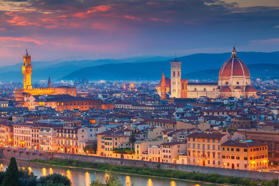 Una città illuminata con una grande cupola e un'alta torre lungo un fiume al tramonto, con montagne sullo sfondo.