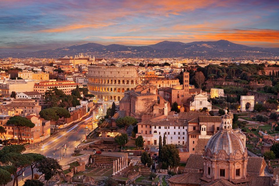 Veduta aerea del Colosseo e del panorama urbano circostante al tramonto, con montagne all'orizzonte sotto un cielo colorato.