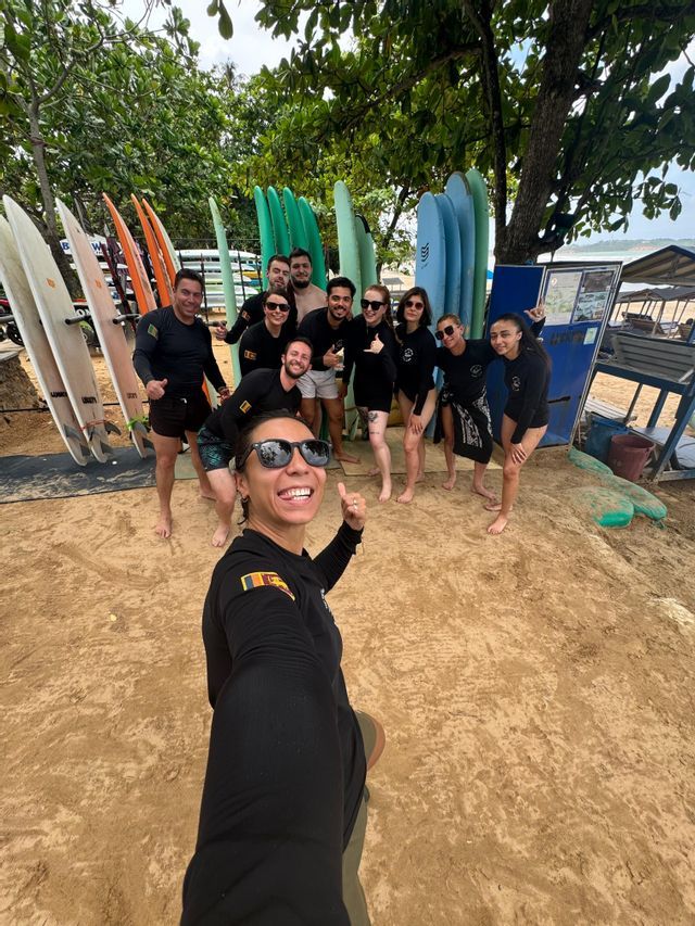 Un groupe WeRoad se prend en selfie sur une plage de sable devant des râteliers de planches de surf colorées.