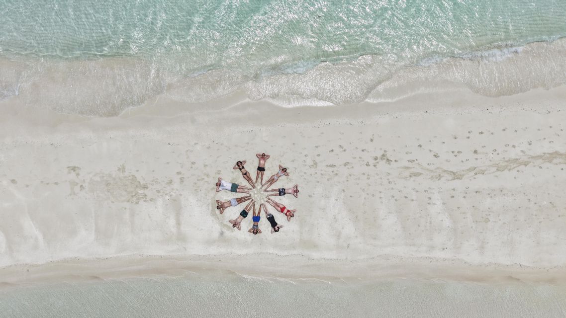 Vue aérienne d'un voyage en groupe WeRoad allongé en cercle sur une plage de sable blanc à côté d'une eau turquoise claire.