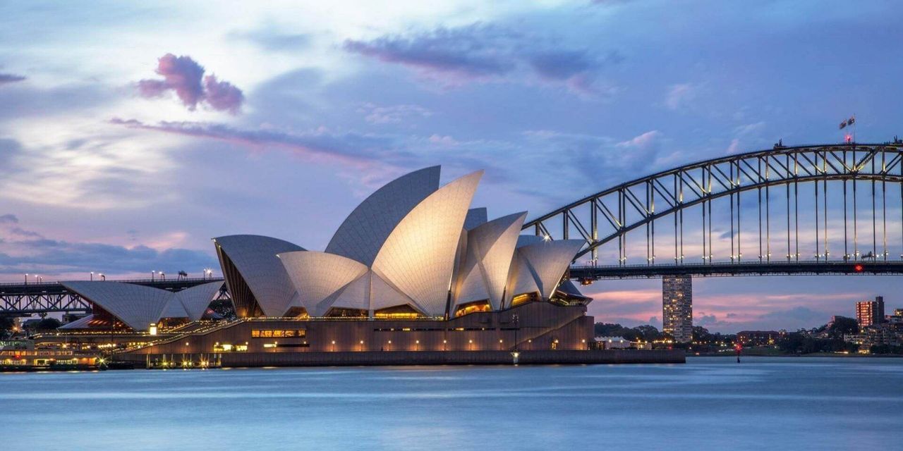 Un edificio moderno con tetti bianchi a forma di vela si erge illuminato accanto a un grande ponte ad arco sull'acqua al tramonto.