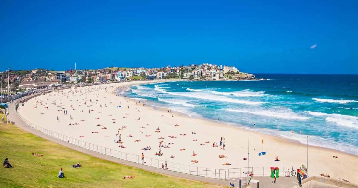 Une vue plongeante d'une plage de sable bondée par une journée ensoleillée, avec des gens qui se baignent dans l'océan et bronzent sur le rivage.