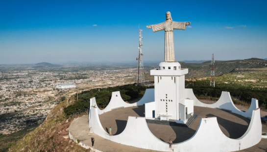 Una veduta aerea di una grande statua di Cristo, posta su una piattaforma bianca in cima a una collina, che domina un'ampia città sotto un cielo blu.