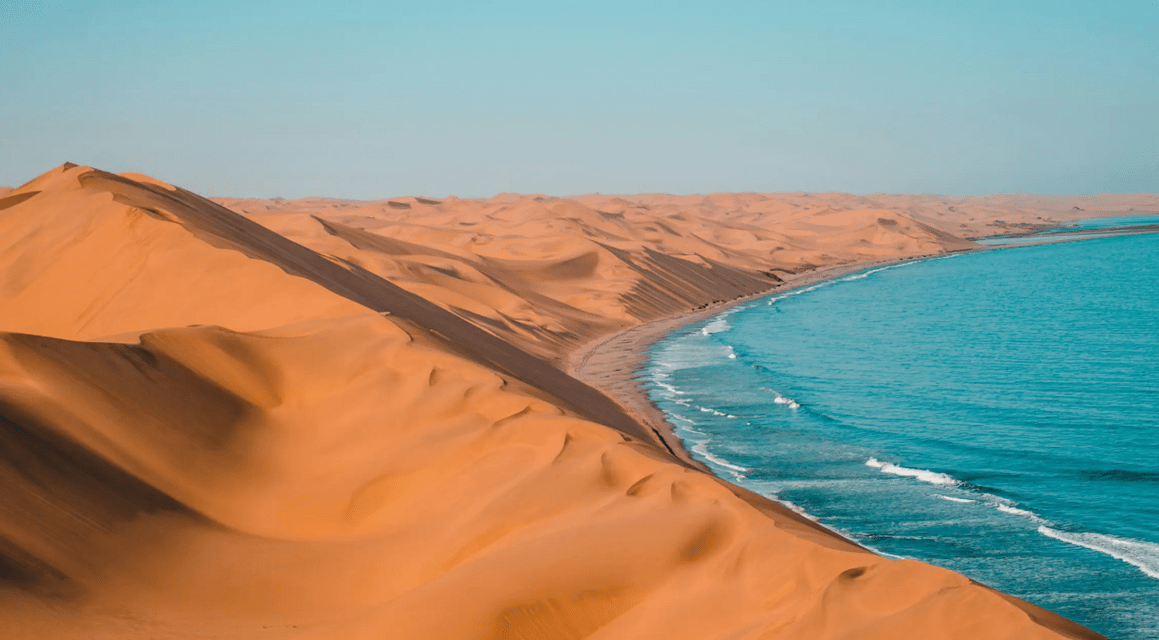 Vaste dune di sabbia arancione incontrano l'oceano blu lungo una costa sotto un cielo sereno.