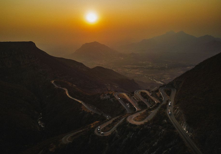Veduta aerea di una strada tortuosa con tornanti che scende da una montagna, con auto che la percorrono durante un tramonto velato.