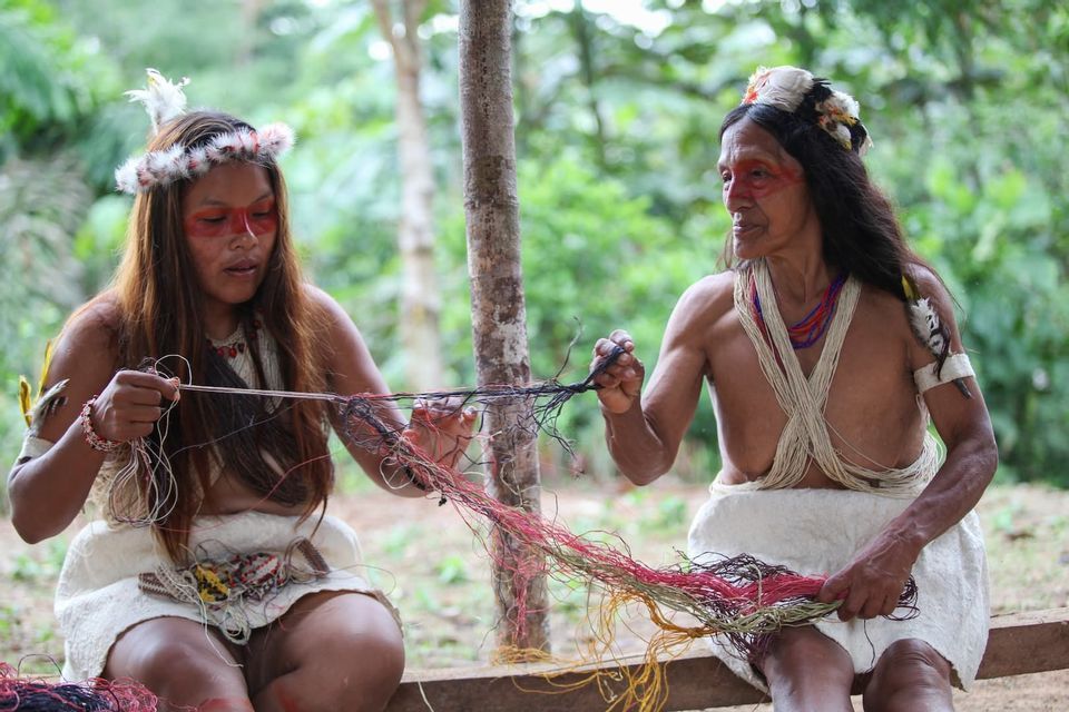 Dos mujeres indígenas con ropa tradicional y tocados de plumas trabajan juntas con hilos coloridos al aire libre