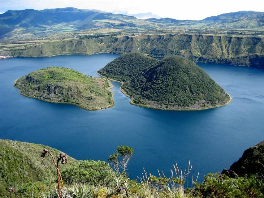 Una vista dall'alto di due isole verdi in un lago craterico blu intenso, circondato da dolci colline sotto un cielo sereno.