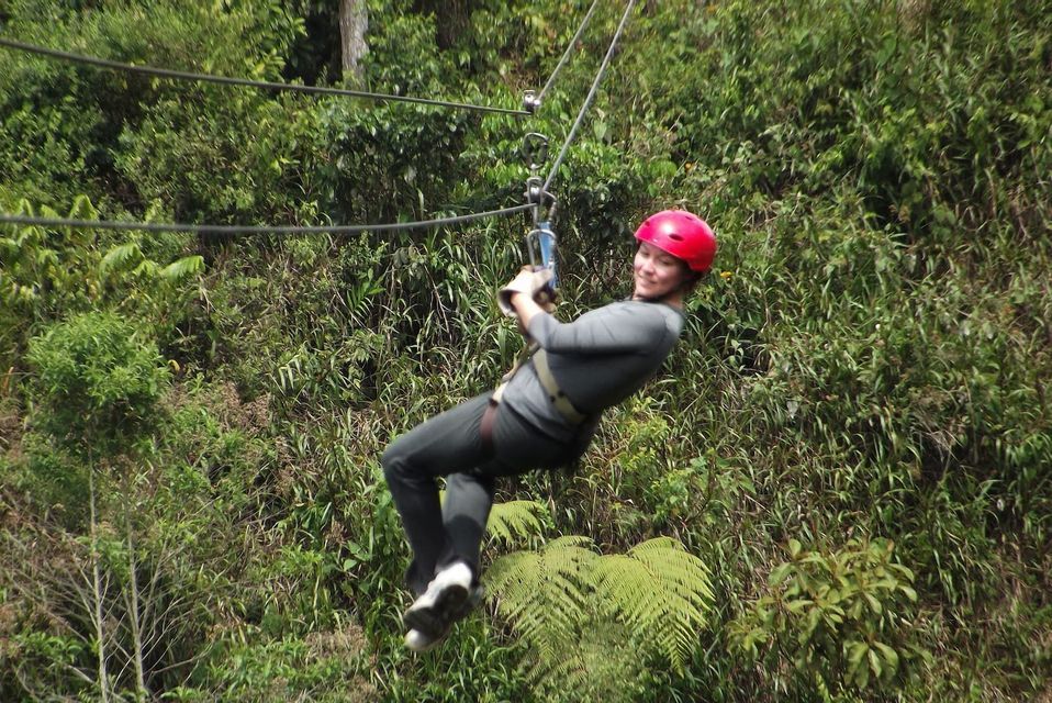 Una persona con casco rosso sorride mentre fa zipline in una fitta foresta verde.