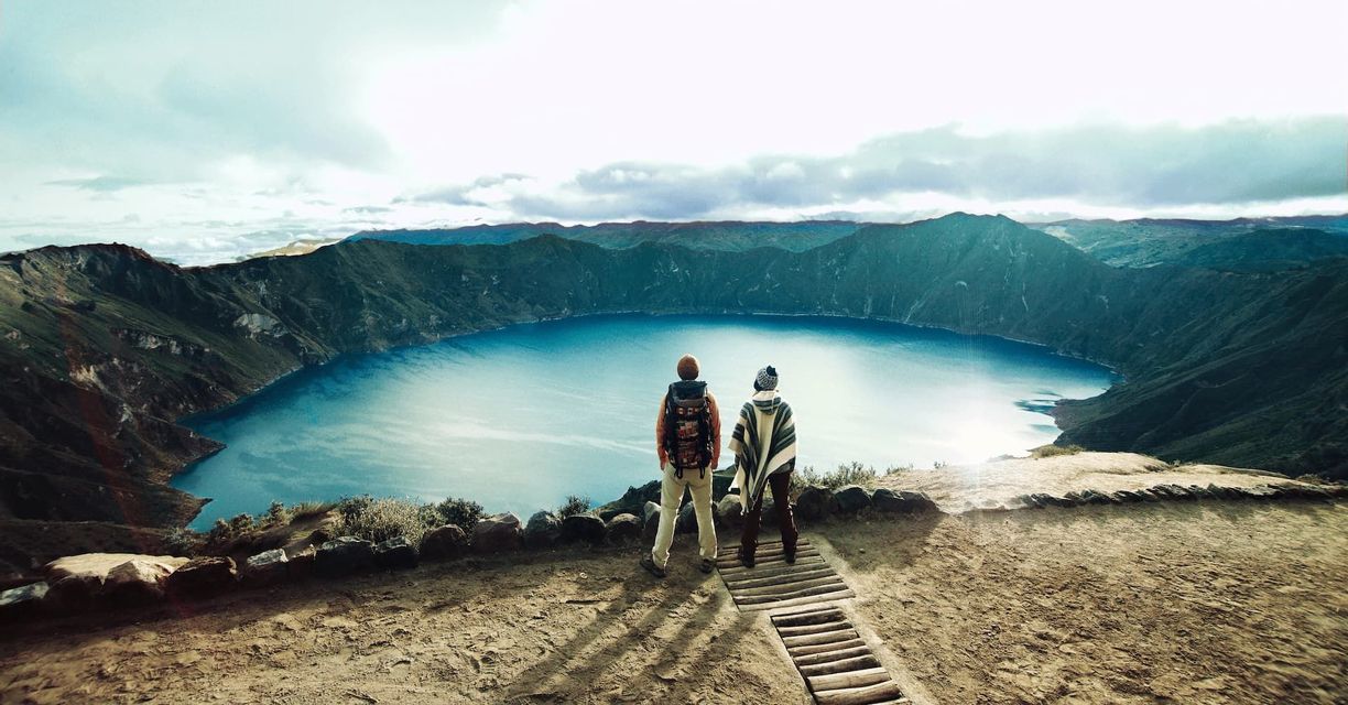 Due persone viste di spalle, una con uno zaino e l'altra con un poncho, si trovano su un punto panoramico che si affaccia su un lago vulcanico.