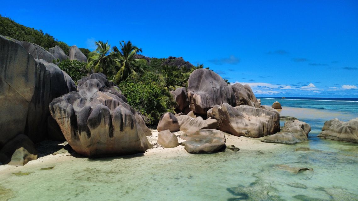 Enormi massi erosi si adagiano su una spiaggia sabbiosa bagnata da acque limpide e turchesi, con palme e una vegetazione lussureggiante su una collina sullo sfondo.