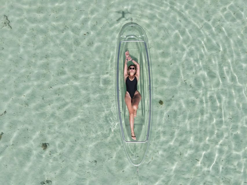 Vista dall'alto di una donna in costume nero sdraiata in un kayak trasparente che galleggia su acqua limpida e poco profonda.