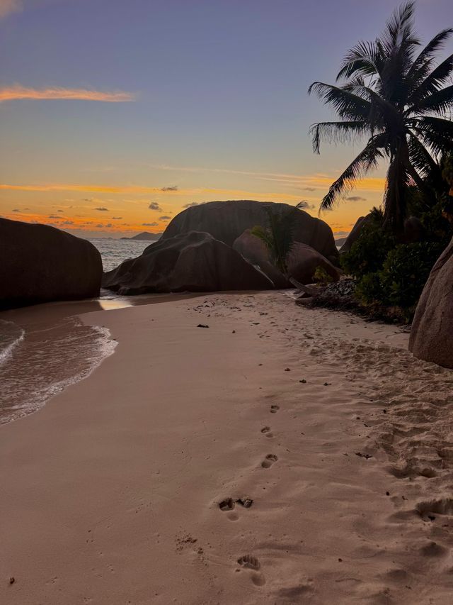Impronte nella sabbia su una spiaggia tropicale con grandi massi e una palma durante un tramonto colorato.