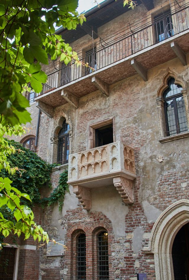 Un balcone in pietra ornato si estende da un edificio in mattoni anticato con finestre ad arco, incorniciato da foglie verdi in primo piano.